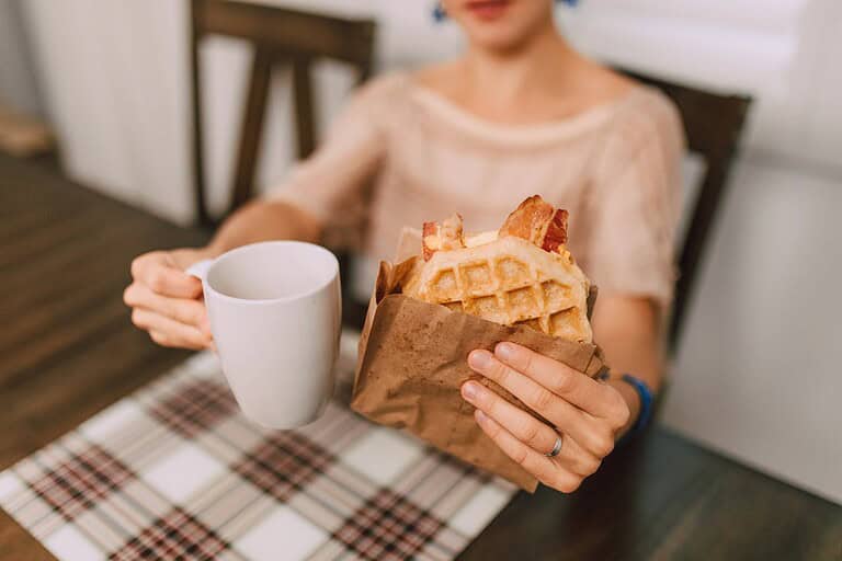A person holding a white mug in one hand and a brown paper bag containing a waffle with bacon in the other
