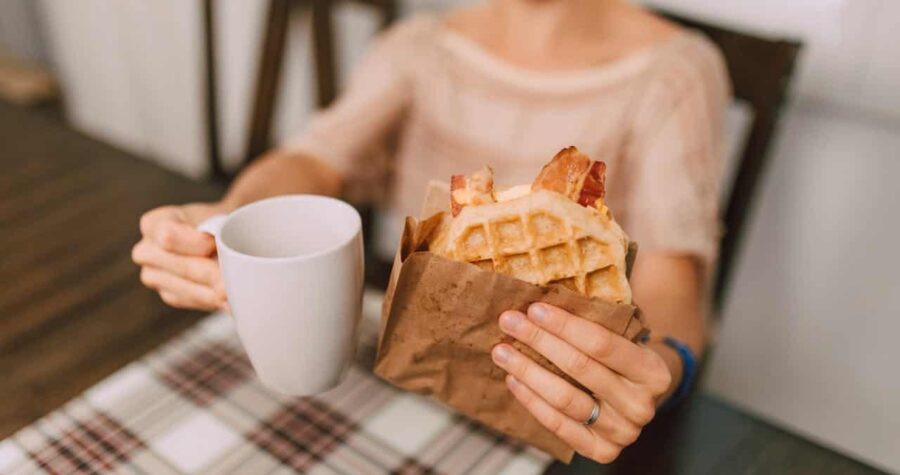 A person holding a white mug in one hand and a brown paper bag containing a waffle with bacon in the other