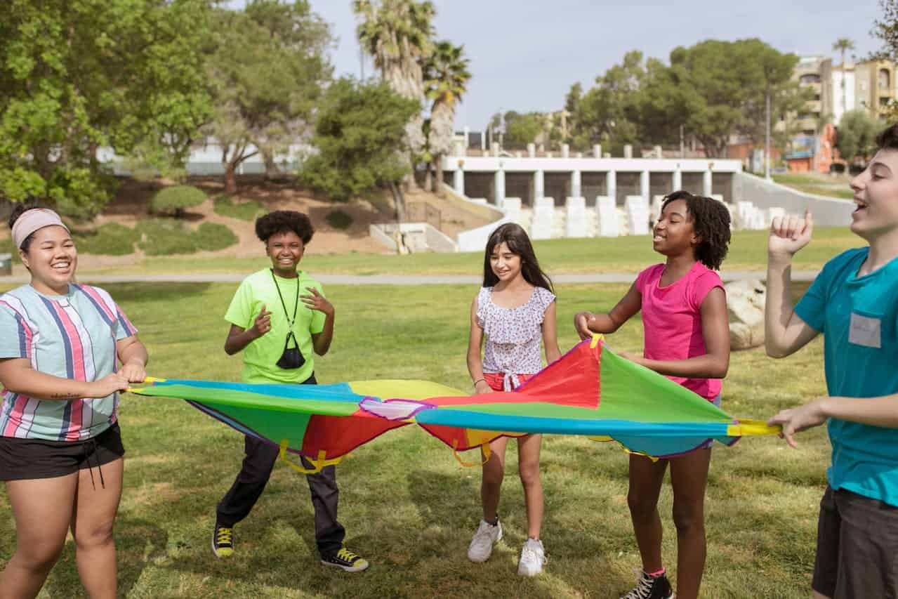 A group of five children stands together on a grassy field, each holding a colorful parachute, smiling and enjoying the moment, the children are engaged in a fun outdoor activity, the bright colors of the parachute create a lively atmosphere, with trees and buildings in the background
