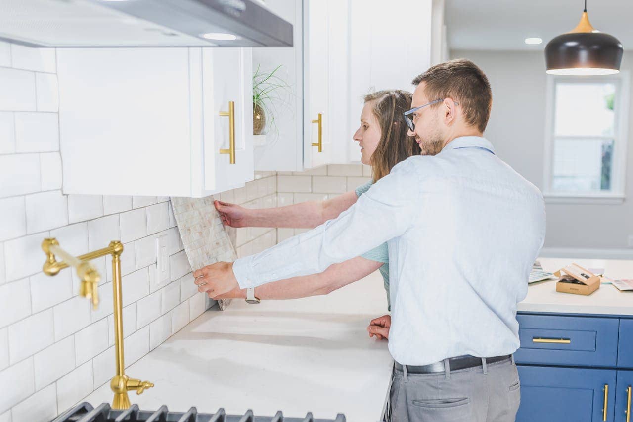 Two people collaborating to install kitchen backsplash tiles, one person holding a tile up against the wall, the other person observing and assisting, modern kitchen design with white cabinets, gold accents, clean countertops, bright lighting from a pendant light, tools and tiles visible on the counter