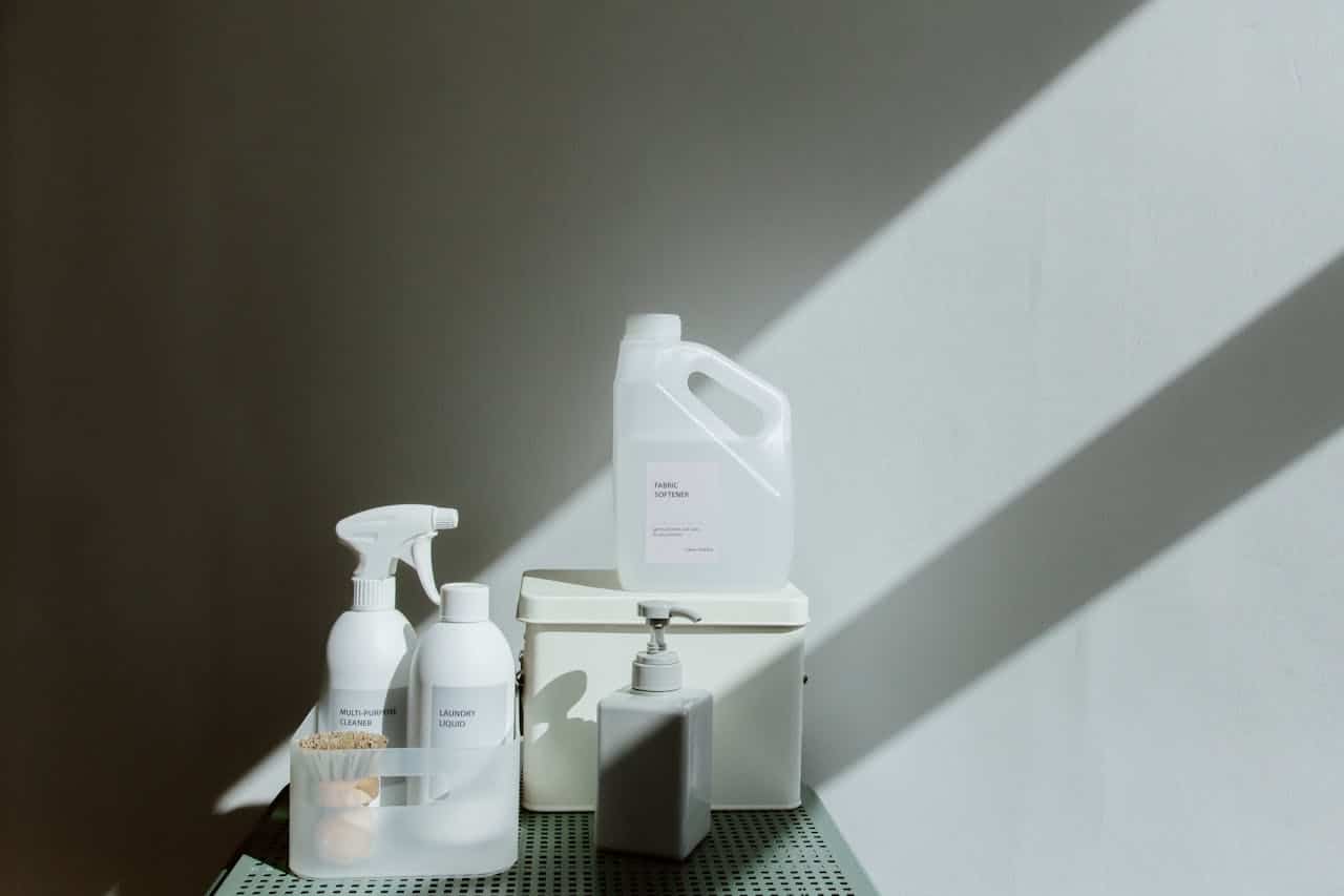 Minimalist white cleaning products including fabric softener, spray cleaner, and laundry liquid arranged on shelf with dramatic sunlight
