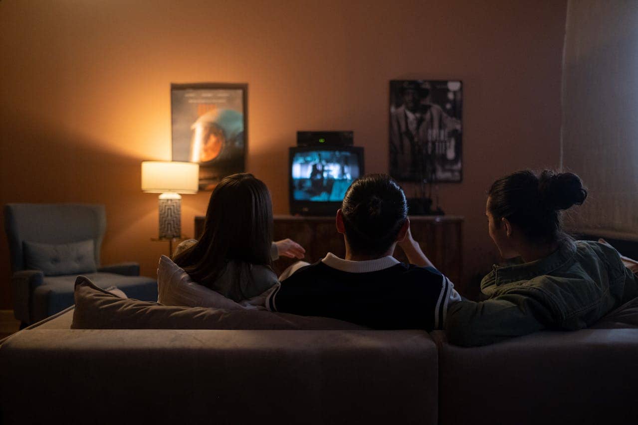 Three people sitting on couch watching TV in dimly lit room with movie posters and lamp visible