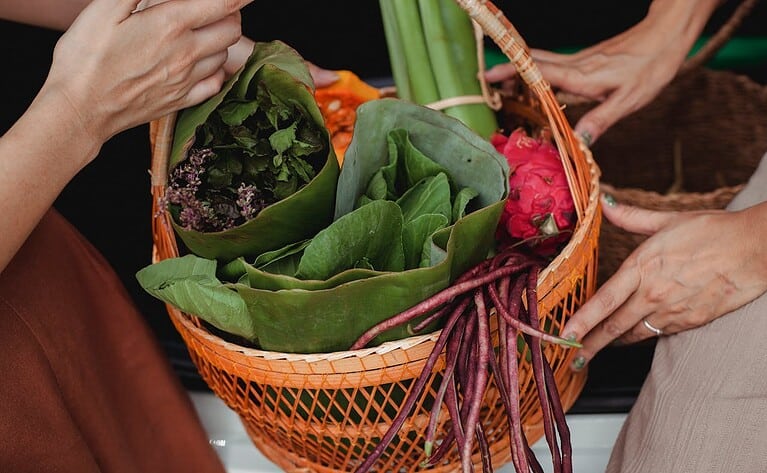 Hands holding orange woven basket filled with fresh produce including leafy greens, purple nasturtiums flowers, dragon fruit, and long purple beans