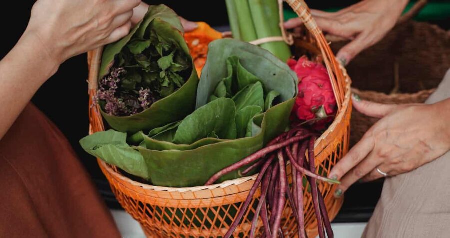 Hands holding orange woven basket filled with fresh produce including leafy greens, purple nasturtiums flowers, dragon fruit, and long purple beans