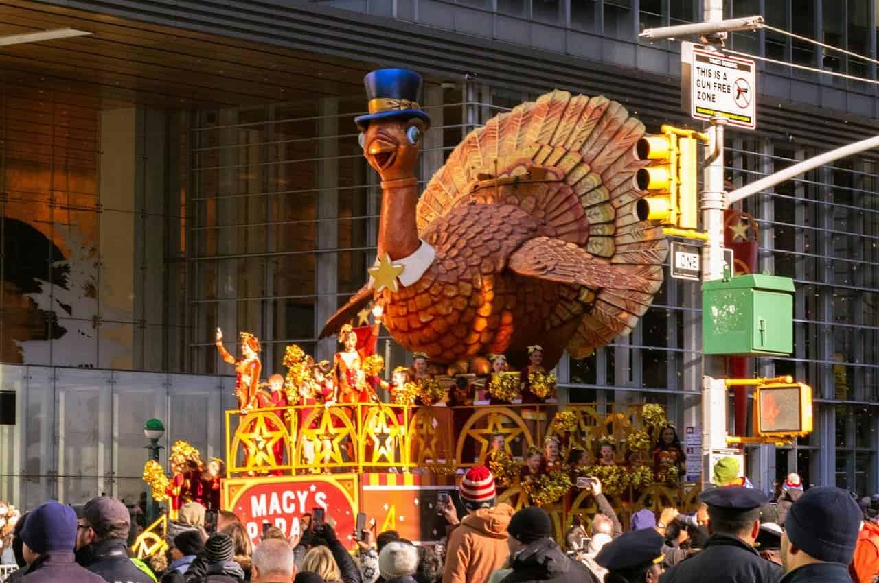 A large Thanksgiving parade float featuring a giant turkey with a blue hat, surrounded by performers in red costumes holding pom-poms, people in the crowd taking photos, a Macy's Parade sign on the float, a busy city street with traffic lights, people enjoying the parade, festive atmosphere