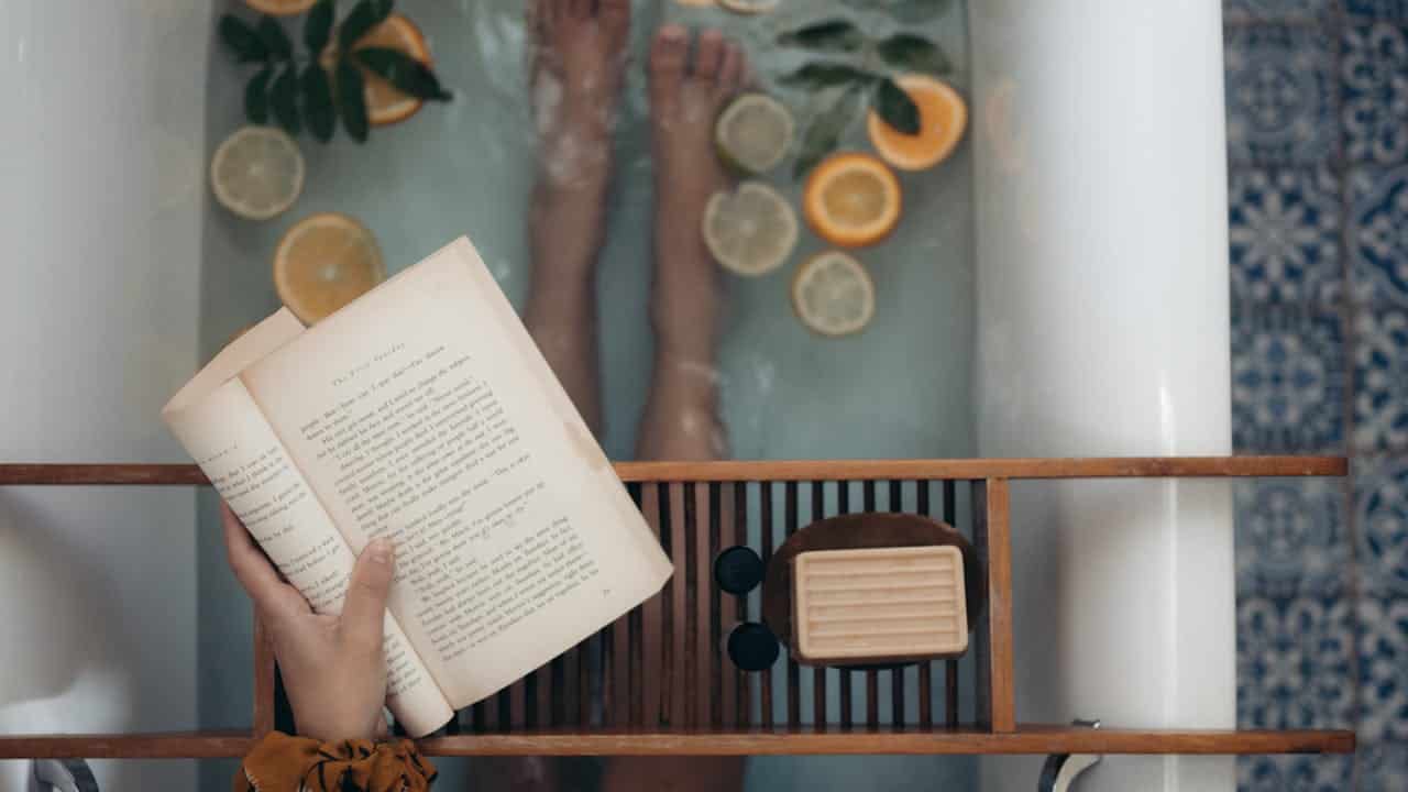 Person reading book while soaking in bathtub with citrus slices, green leaves, and wooden bath tray visible