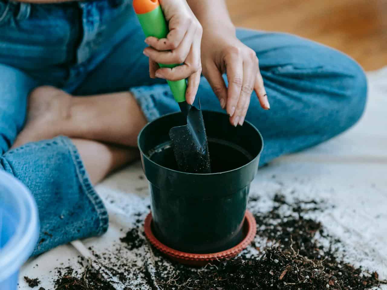 A person sitting on the floor, wearing denim jeans, holding a small green-handled trowel, filling a black plant pot with soil, scattered soil around the pot, the background blurred, preparing for planting