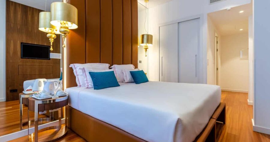 Bedroom with king bed, brown upholstered headboard, gold light fixtures, teal accent pillows, and wooden flooring