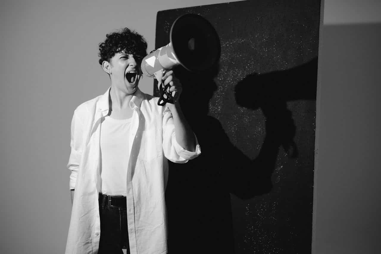 A person shouting into a megaphone, wearing a white shirt and jeans, with curly hair, the shadow of the megaphone clearly visible on the wall behind them, black and white photo emphasizing the action and intensity