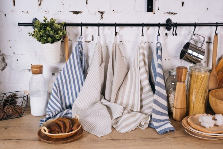 Striped kitchen towels hanging neatly on a black rod against a white brick wall, hung to dry, surrounded by kitchen items like jars of pasta, plates, milk bottle, and a potted plant on a wooden countertop