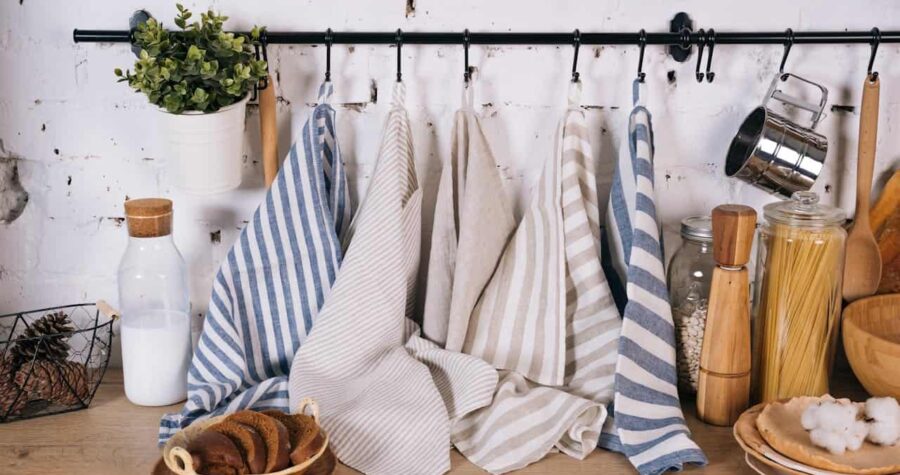 Striped kitchen towels hanging neatly on a black rod against a white brick wall, hung to dry, surrounded by kitchen items like jars of pasta, plates, milk bottle, and a potted plant on a wooden countertop