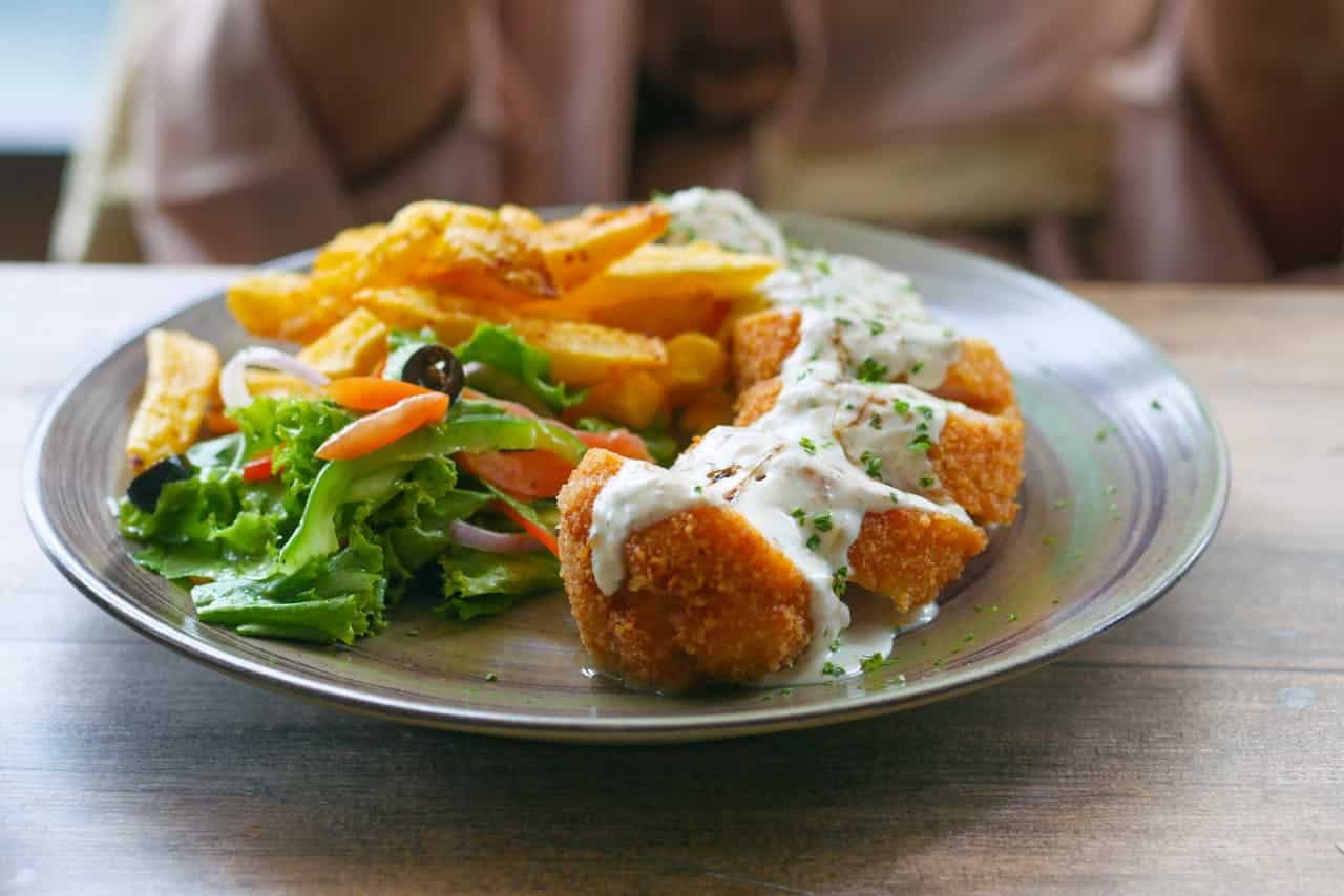 Chicken Cordon Bleu served with golden fried potato wedges, fresh salad with lettuce, carrots, and olives, topped with creamy sauce, garnished with chopped herbs, all on a round plate with a wooden background