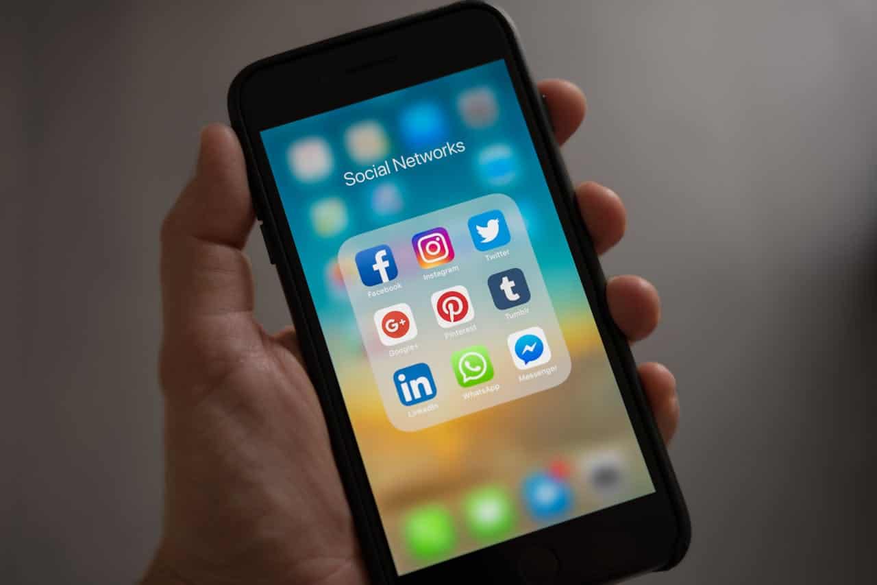 Hand holding smartphone displaying social media folder with Facebook, Instagram, Twitter, Pinterest, WhatsApp and other app icons