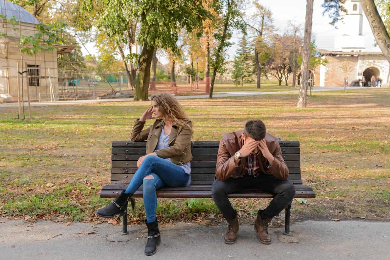 A couple sitting apart on a park bench, looking distressed, the woman touches her head with her hand, the man has his head in his hands, they appear to be in the middle of a tense moment or argument