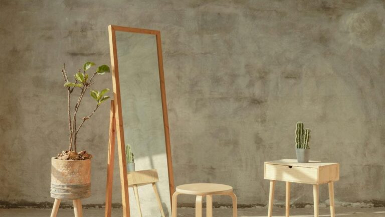 Wood-framed full-length mirror leaning against a textured wall, accompanied by a round wooden stool, a small table, and a potted plant, arranged in a minimalist interior setting with natural lighting