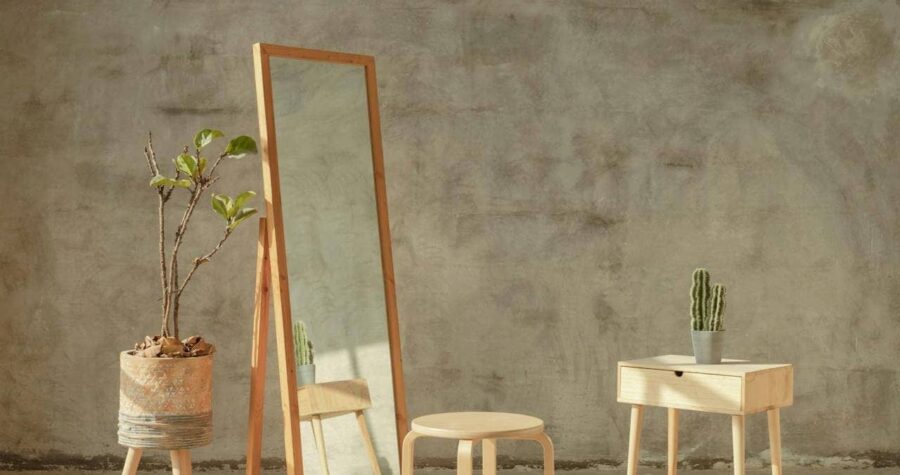 Wood-framed full-length mirror leaning against a textured wall, accompanied by a round wooden stool, a small table, and a potted plant, arranged in a minimalist interior setting with natural lighting