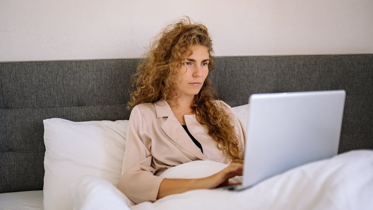 Person with curly hair in light blazer working on laptop while sitting in bed against gray headboard