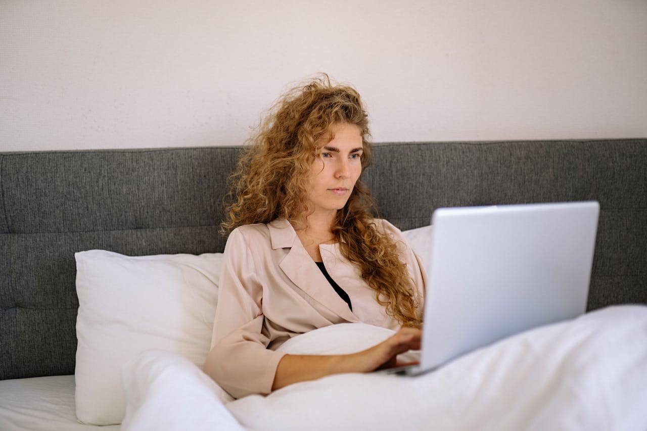 Person with curly hair in light blazer working on laptop while sitting in bed against gray headboard