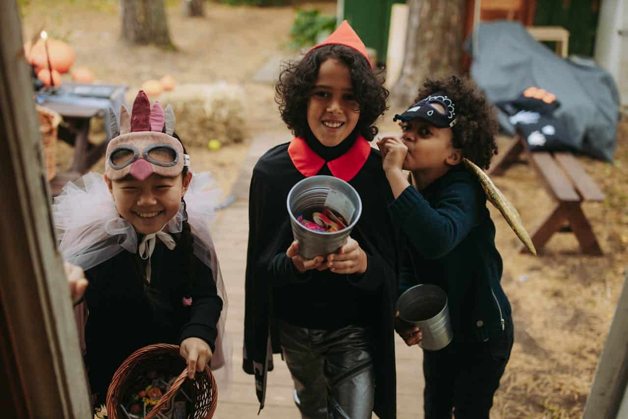 Children dressed in Halloween costumes smiling, one child in a bird costume holding a basket filled with candy, another in a vampire cape holding a bucket, a third child with a bat mask eating candy, background with pumpkins and autumn decorations