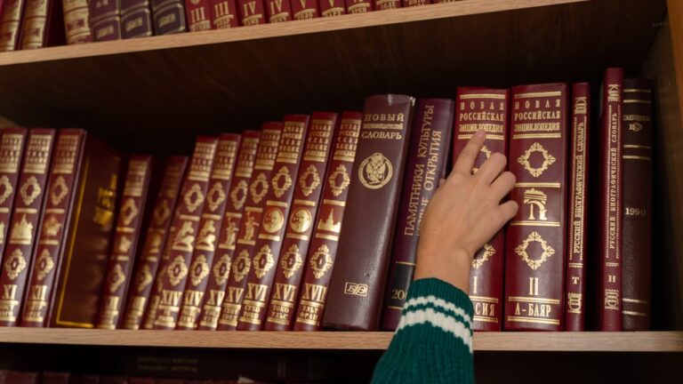 A hand reaching for a book on a shelf filled with leather-bound volumes, the books are titled in gold lettering, and the shelf contains various encyclopedias and reference books