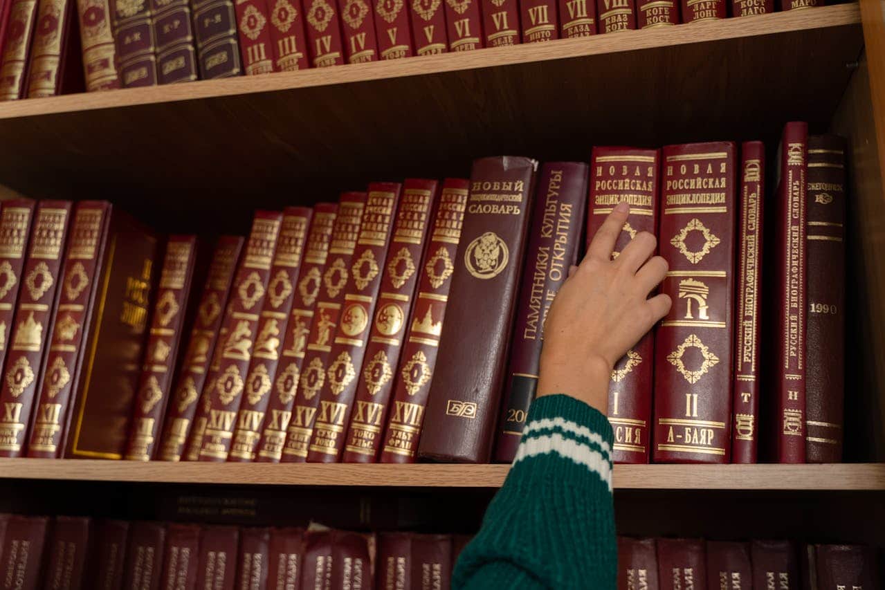 A hand reaching for a book on a shelf filled with leather-bound volumes, the books are titled in gold lettering, and the shelf contains various encyclopedias and reference books