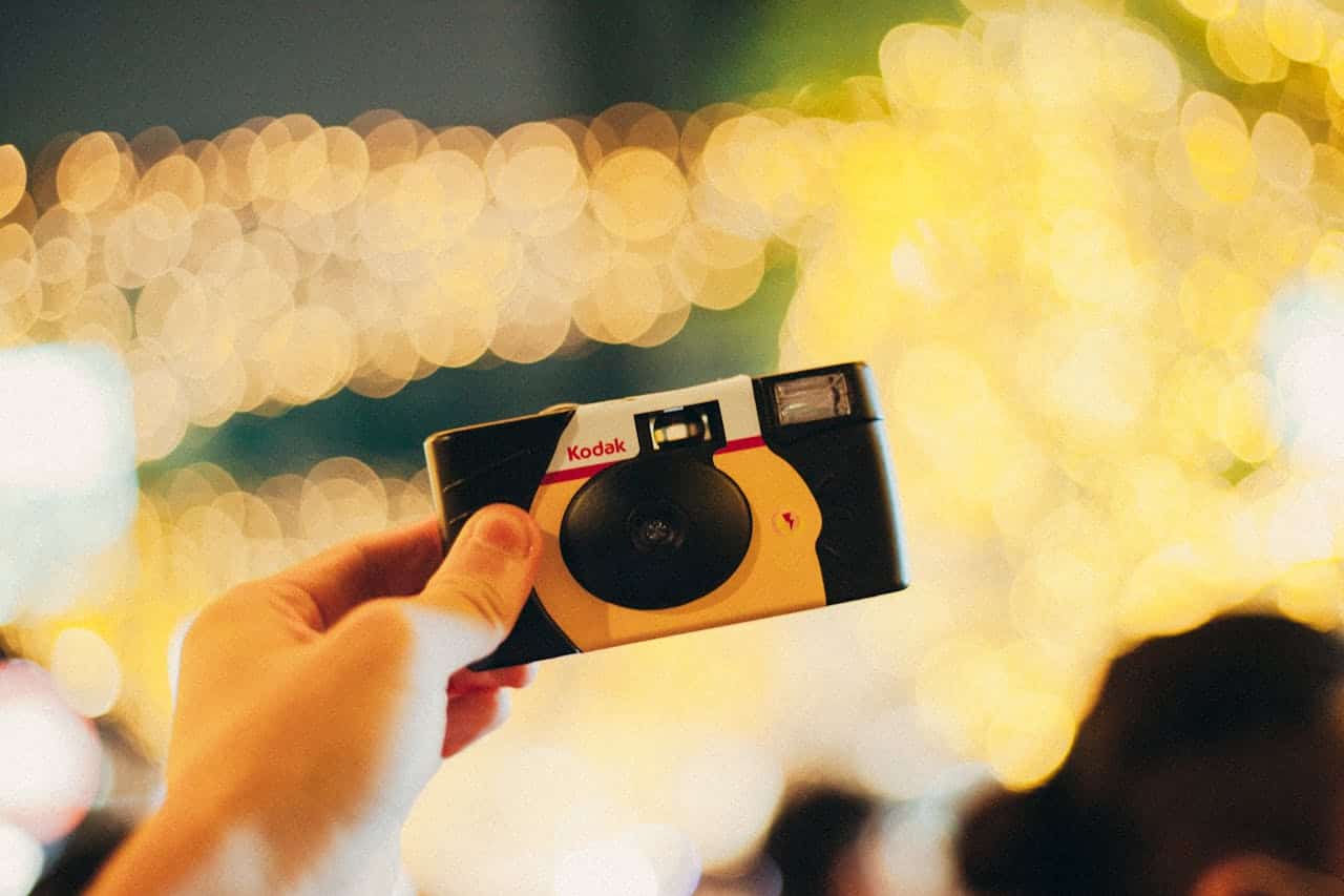 A person holding a Kodak camera in front of a blurred background with soft bokeh lights, the camera is black and yellow with a flash, capturing a vintage, nostalgic feel