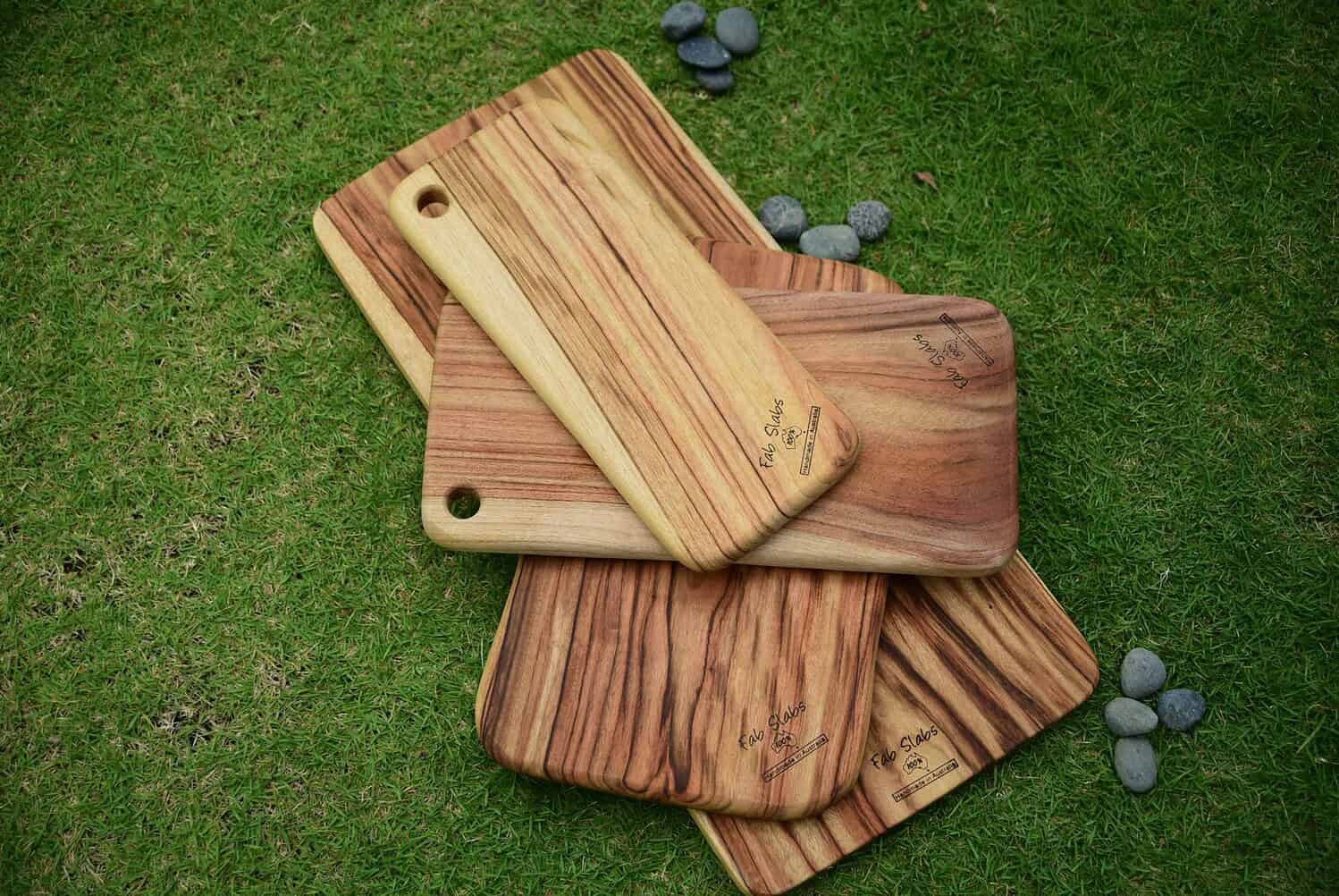 Wooden cutting boards with a striped pattern stacked on grass, each board has a hole for hanging, smooth finish, and engraved logos on the surface, natural earthy tones of the wood contrasting with the vibrant green grass, minimalistic design, aesthetic for kitchen use or display