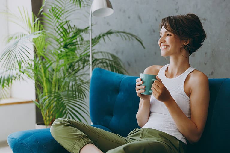 A person sitting on a blue couch, holding a cup of tea, smiling and relaxed, surrounded by a potted plant and natural light, wearing casual clothes, creating a peaceful, cozy atmosphere of contentment and calm