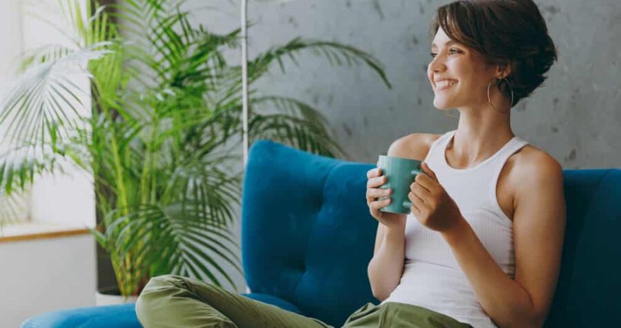 A person sitting on a blue couch, holding a cup of tea, smiling and relaxed, surrounded by a potted plant and natural light, wearing casual clothes, creating a peaceful, cozy atmosphere of contentment and calm