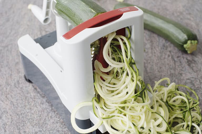 White spiralizer kitchen tool creating zucchini noodles from fresh zucchini on a marble countertop surface