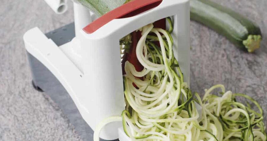 White spiralizer kitchen tool creating zucchini noodles from fresh zucchini on a marble countertop surface