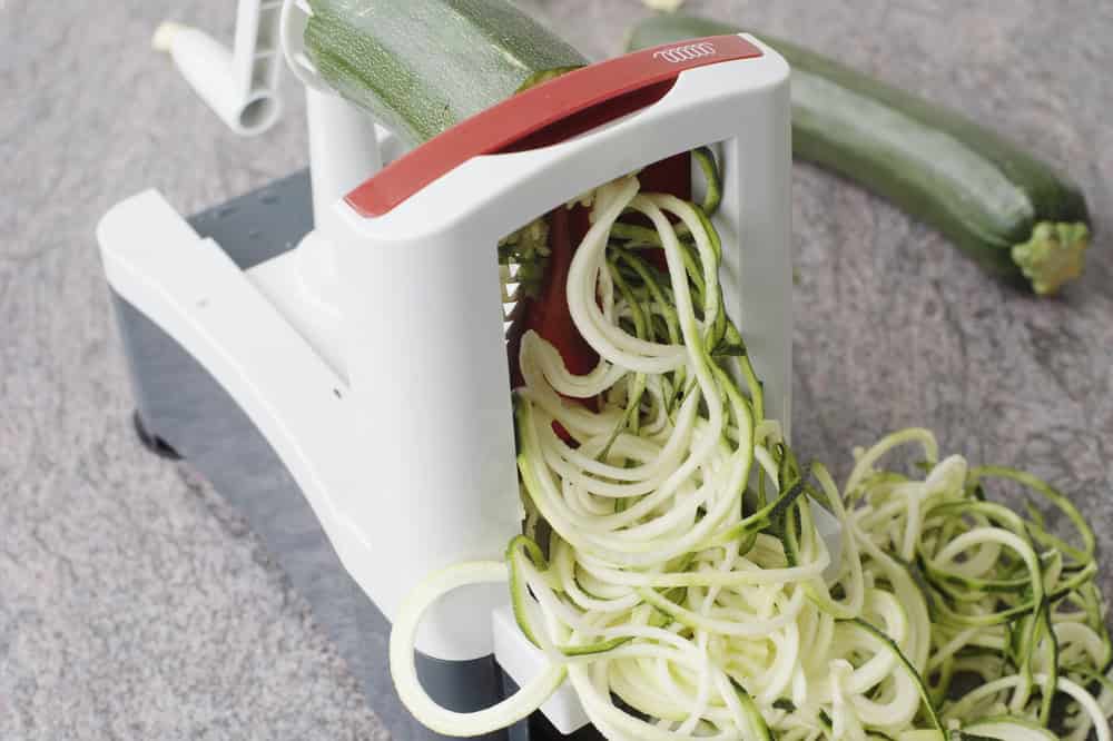 White spiralizer kitchen tool creating zucchini noodles from fresh zucchini on a marble countertop surface