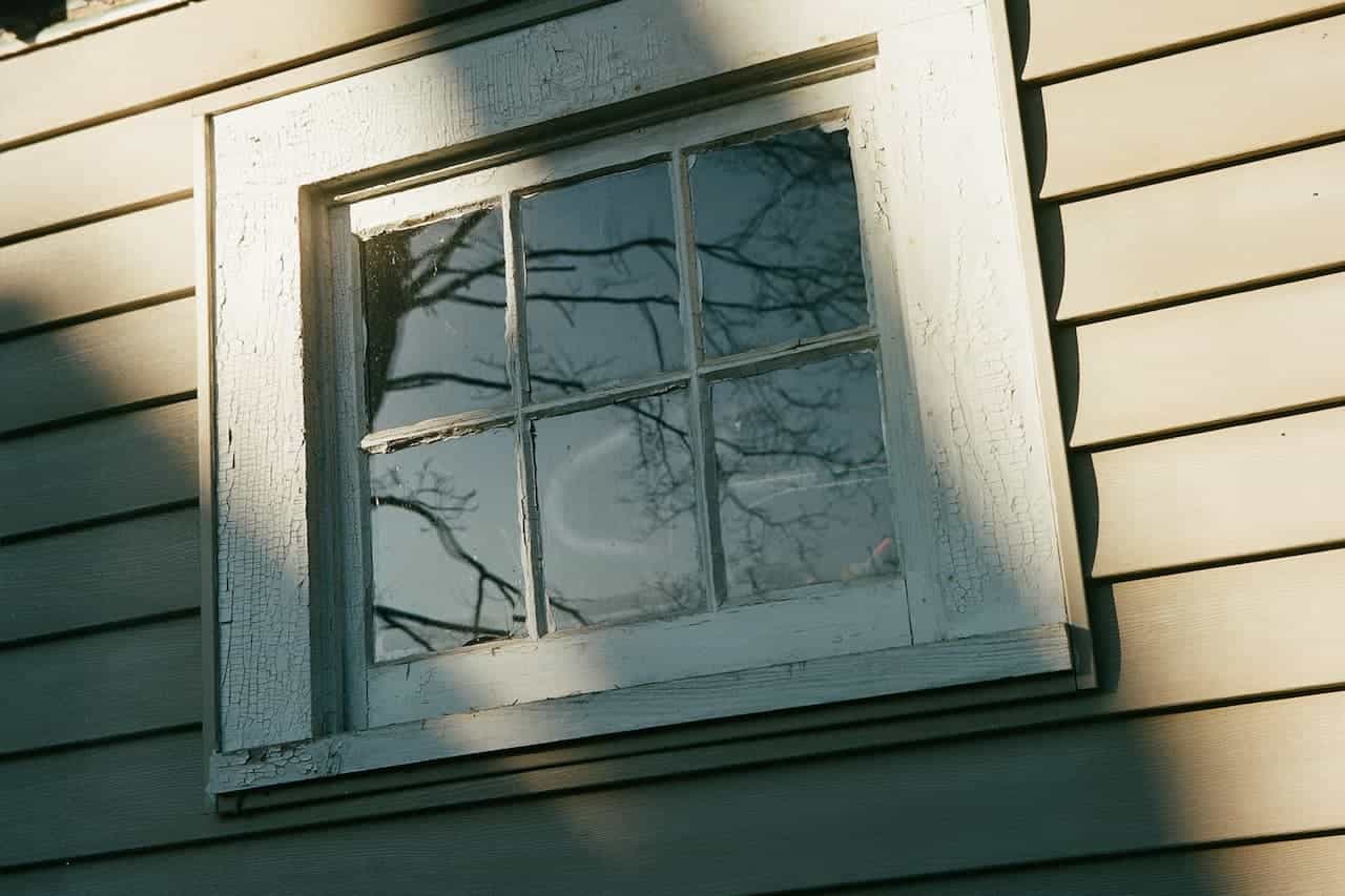 Window with six panes in white wooden frame on light-colored siding, reflecting bare tree branches against blue sky