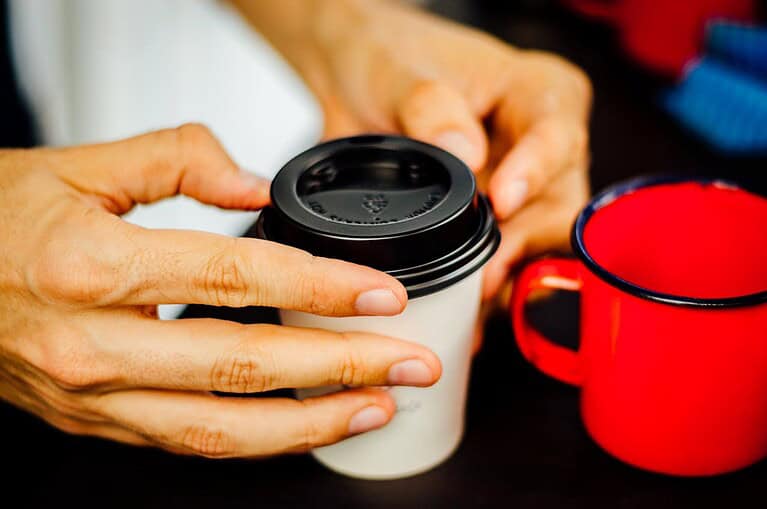 Hands holding a white disposable coffee cup with a black lid, red ceramic mug nearby, dark surface background, close-up view
