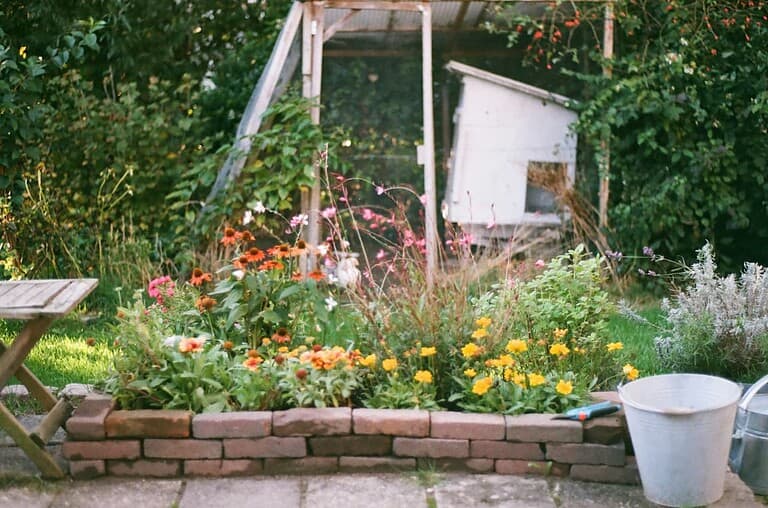 A vibrant garden bed filled with blooming flowers in various colors, a stone wall surrounding the garden, a wooden table placed nearby, a bucket with a gardening tool beside it, a rustic shed in the background, green foliage and trees surrounding the area, peaceful outdoor setting