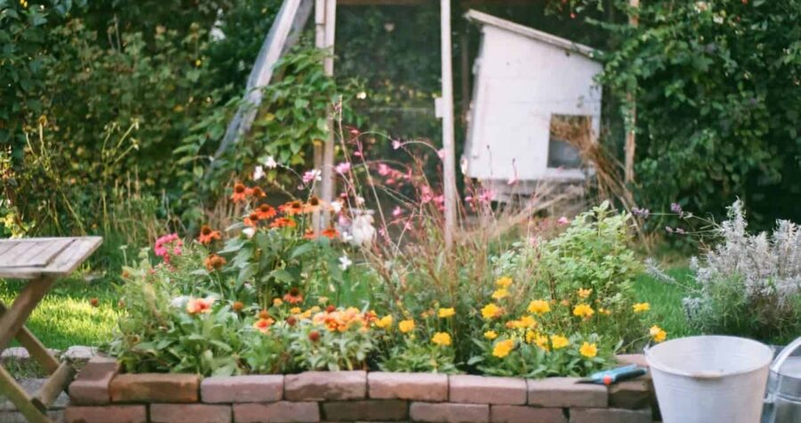 A vibrant garden bed filled with blooming flowers in various colors, a stone wall surrounding the garden, a wooden table placed nearby, a bucket with a gardening tool beside it, a rustic shed in the background, green foliage and trees surrounding the area, peaceful outdoor setting