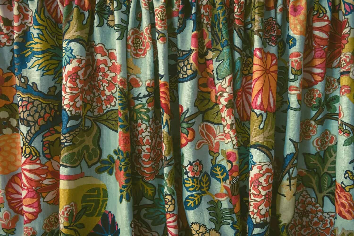 The image shows a close-up view of vibrant, floral patterned curtains, with colorful flowers like reds, oranges, and pinks, contrasting against a soft blue background, the fabric is gently folded, adding a textured look to the scene