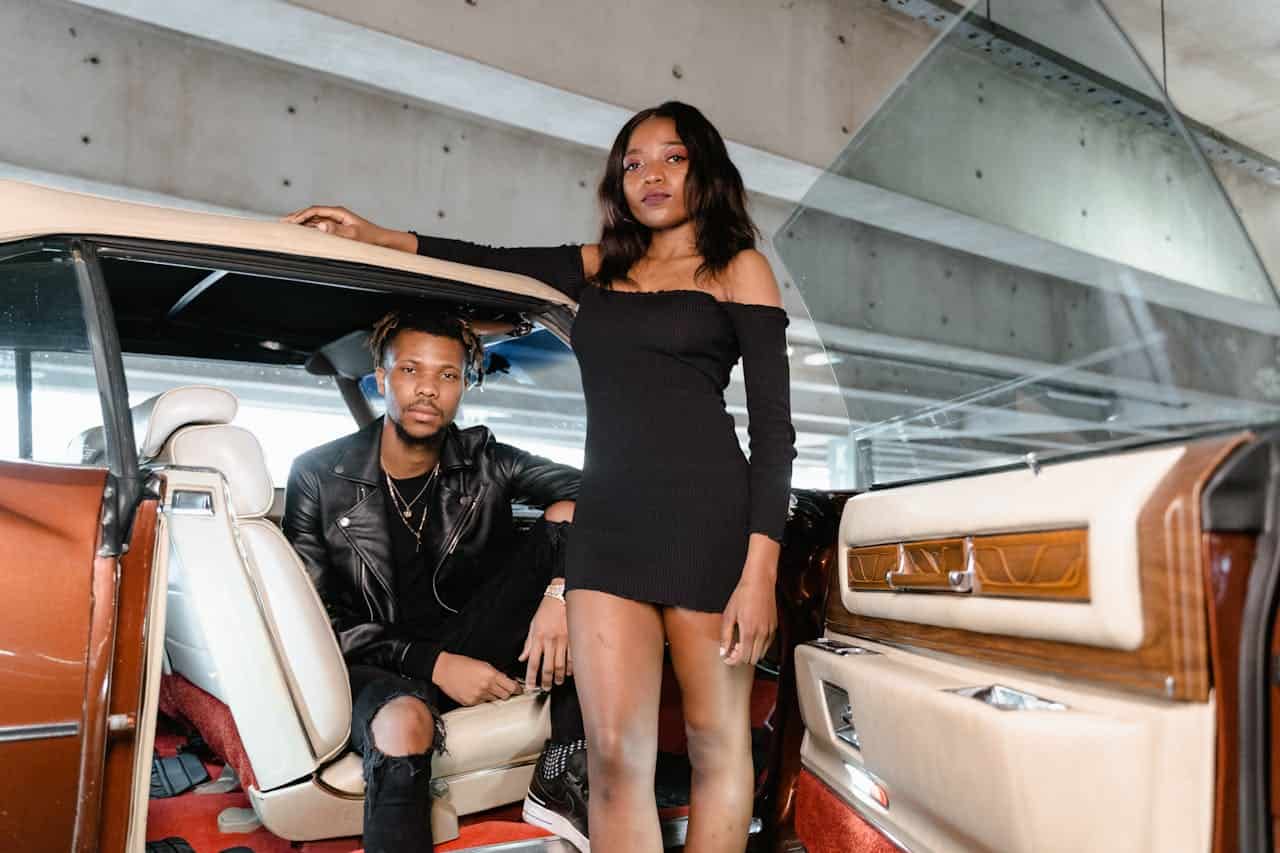 Two young people in a vintage luxury car with cream leather seats and wood paneling. A woman in a black dress stands at the door while a man in a leather jacket sits inside