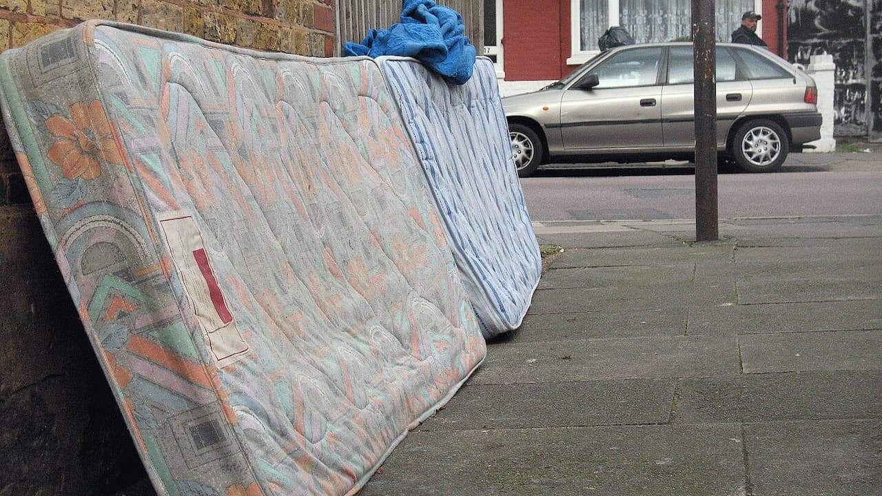 Used mattress with floral pattern and blue blanket leaning against brick wall on sidewalk near parked car