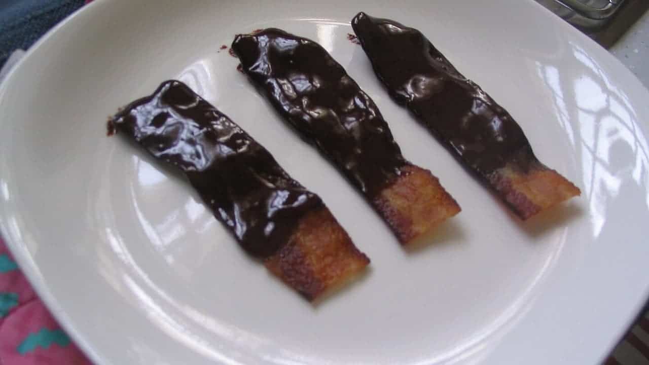Three bacon strips partially dipped in chocolate arranged parallel on a white plate with crispy ends visible