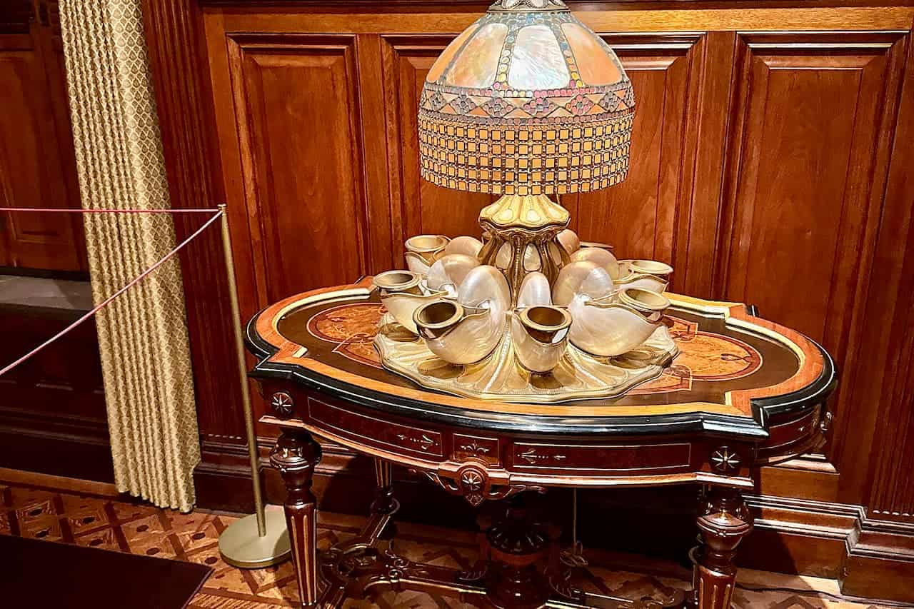 Ornate antique table with inlaid design displaying nautilus shell floor lamp with stained glass shade against wooden paneled wall