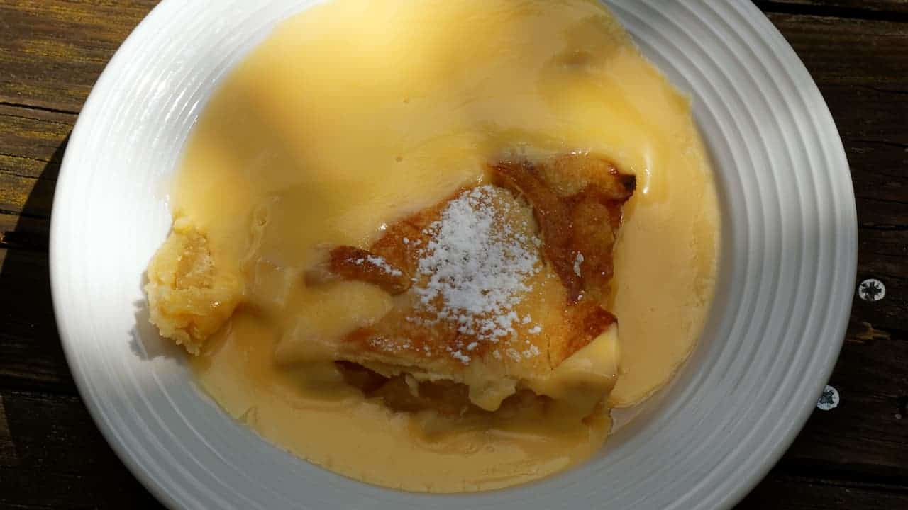 Apple dessert with custard sauce on white plate, dusted with powdered sugar, served on dark wooden table