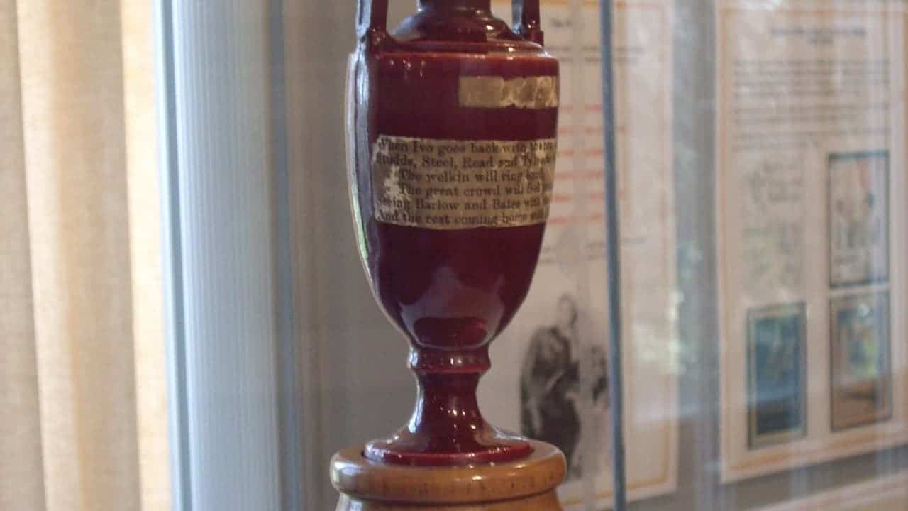 Dark red ceramic memorial urn with gold band and text label displayed on pedestal in museum or memorial setting