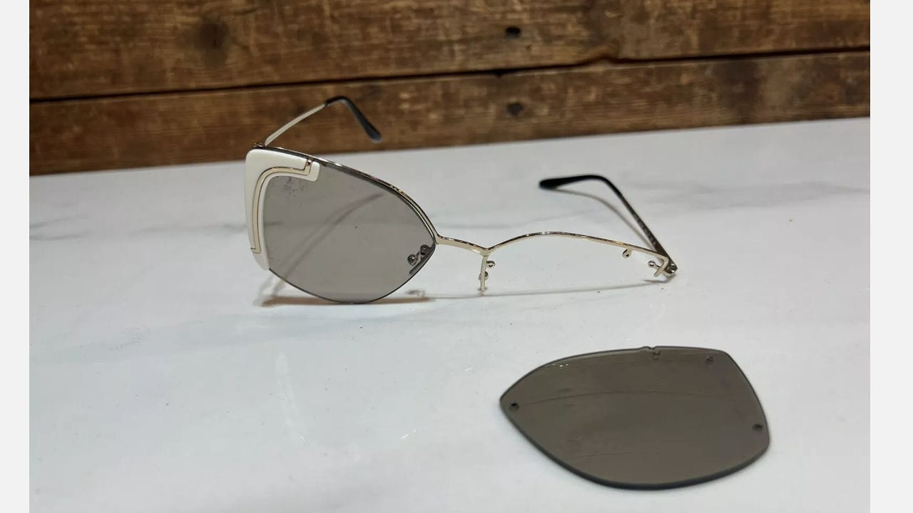 A pair of broken sunglasses with one detached lens lying next to the frame on a white surface.