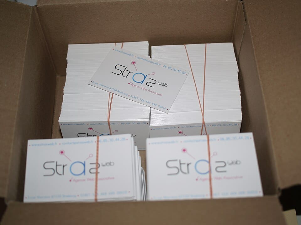Box of stacked business cards, white cards with blue and pink text, rubber bands securing stacks, one card slightly tilted, branding reads "Strasweb", contact details visible.