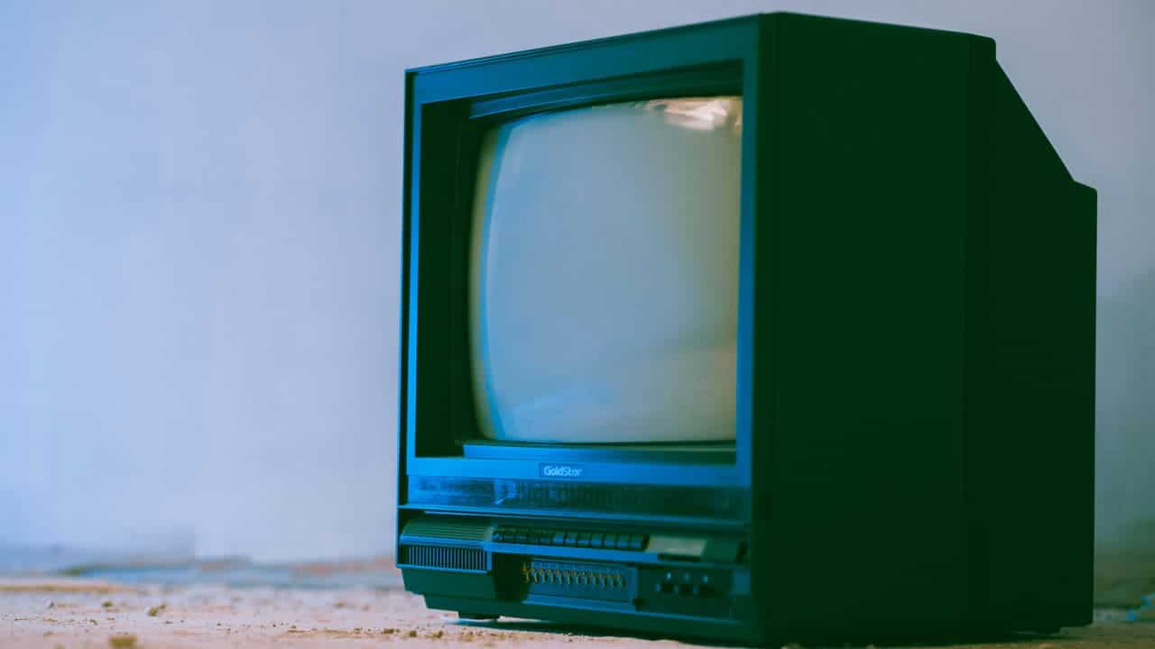 A vintage CRT television with a curved screen, black casing, and manual control buttons, sitting on a rough floor against a plain wall