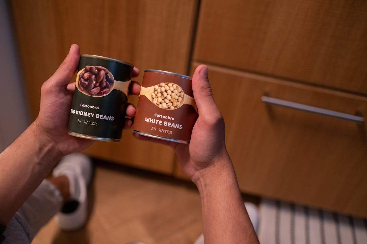 Two hands holding canned red kidney beans and white beans labeled "in water," with wooden cabinets in the background