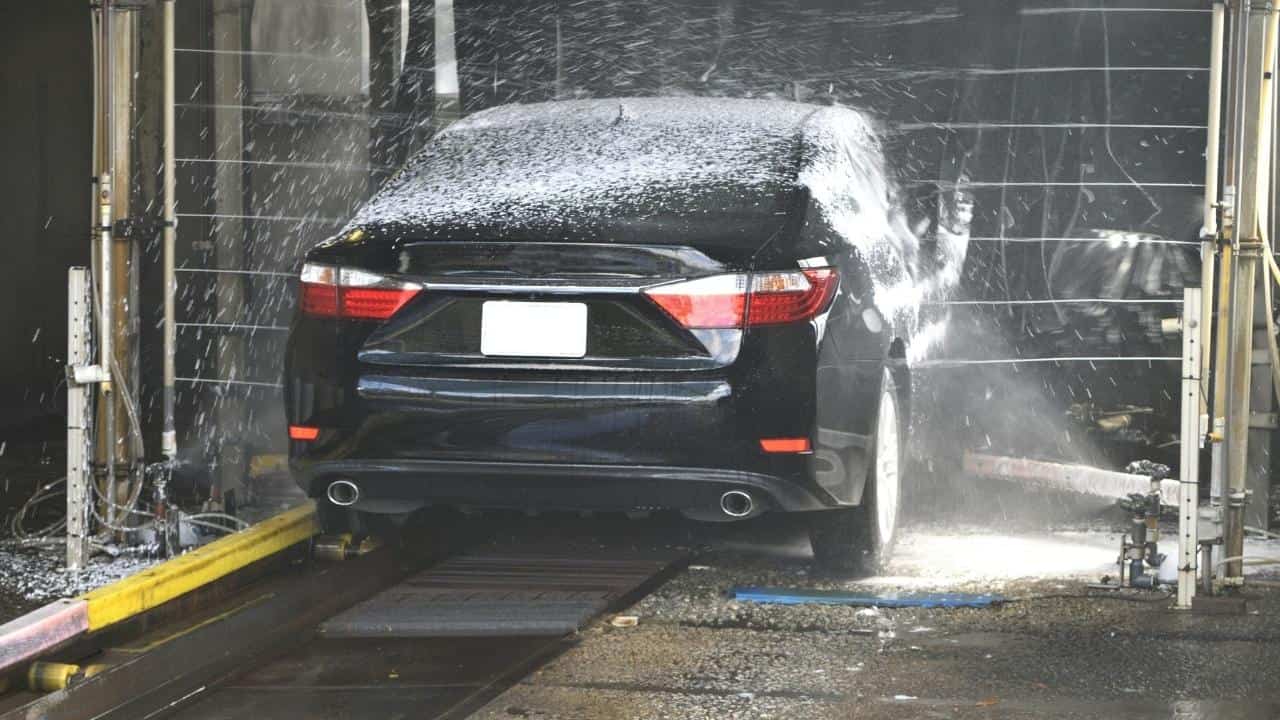 Black sedan going through automated car wash, rear view of vehicle covered in soap and water spray, mechanical wash equipment visible on sides, car positioned on track system