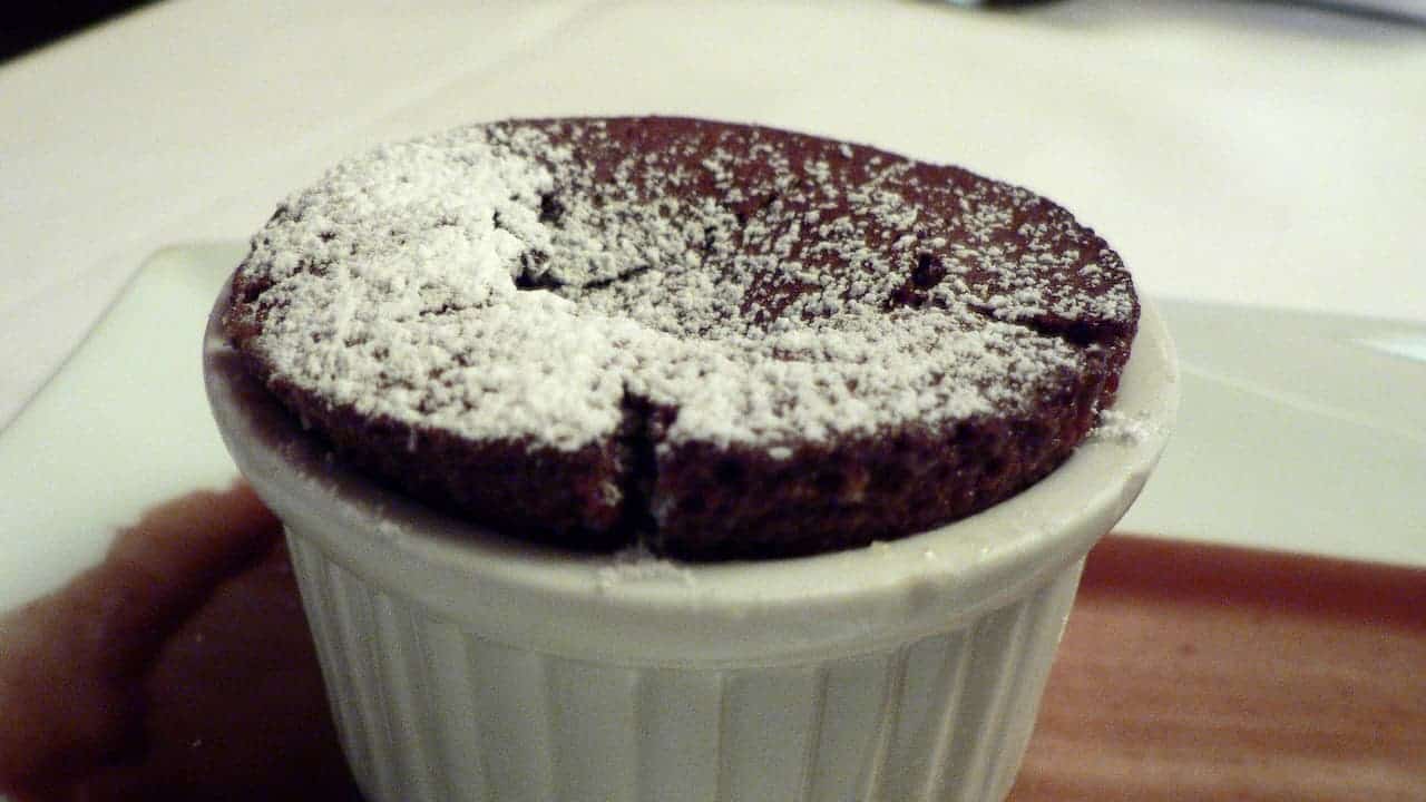 Chocolate soufflé in white ramekin dusted with powdered sugar, showing characteristic crack on top, served on wooden surface