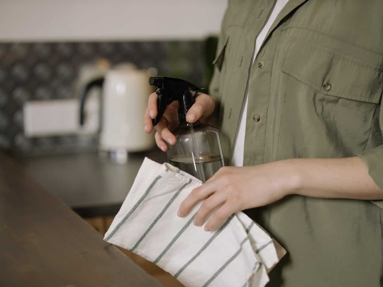 Person holding spray bottle and striped cloth, preparing to clean kitchen counter, wearing green shirt, indoor kitchen setting